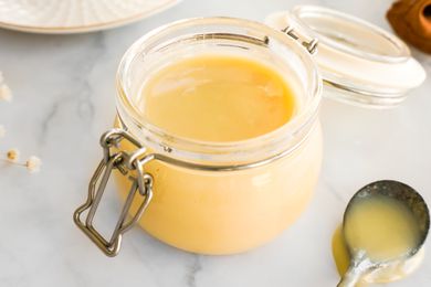 Jar of Homemade Condensed Milk Surrounded by a Spoon With Condensed Milk, a Plate, and a Kitchen Towel